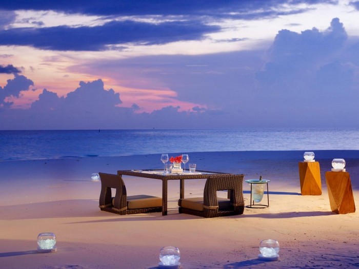 Outdoor-Dining-Table-On-Beach-With-Rattan-Chairs-And-Candle-Glass